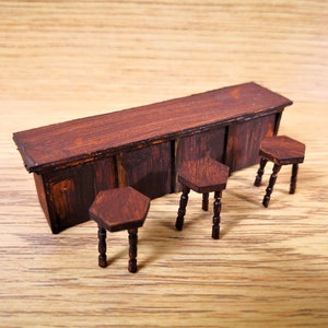 以下が含まれることがあります： 濃い茶色の仕上げが施されたミニチュアの木製バーカウンターと、それに合わせた3つのスツール。バーカウンターには3つのドアと平らな天板があります。スツールは六角形のシートと4本の脚を持っています。