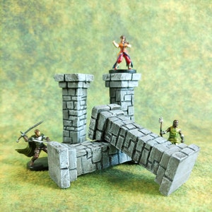 Peut inclure: Quatre piliers en pierre grise, deux debout et deux tombés sur le côté. Les piliers sont texturés pour ressembler à de la pierre brute. Une figurine miniature d'une personne se tient au sommet d'un des piliers debout.