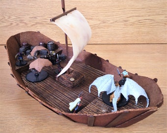 Könnte beinhalten: Ein Miniatur-Holzschiff mit einem weißen Segel, einem braunen Holzdeck und einer weißen Drachenfigur auf dem Deck. Das Schiff ist von Miniaturfiguren von Menschen und Fässern umgeben.