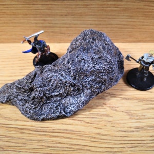 Può includere: Una formazione rocciosa grigia e testurizzata, perfetta per i giochi da tavolo. Due figure in miniatura, una con una spada e l'altra con un'ascia, sono posizionate sulla roccia.