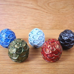 Puede incluir: Cinco huevos de dragón, cada uno con un color y una textura diferentes. Los huevos son azules, blancos, verdes, rojos y negros.
