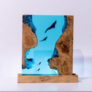 Könnte beinhalten: Ein rechteckiges Holzkunstwerk mit einer blauen Harz-Ozeanszene mit Wal- und Fischsilhouetten. Das Kunstwerk ist von natürlichen Holzkanten umrahmt und steht auf einem Holzsockel, wodurch ein dreidimensionaler Effekt entsteht.