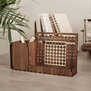 Könnte beinhalten: Ein Schreibtisch-Organizer aus Holz mit Kalenderfunktion. Der Organizer hat Fächer für Stifte und einen Kalender mit den Monaten, die oben beschriftet sind. Der Kalender zeigt die Tage des Monats in einem Raster an. Das Holz hat einen warmen, braunen Ton.