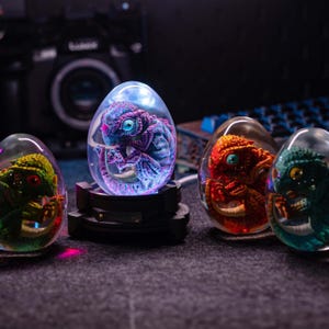 Op de afbeelding: Vier verlichte hars sculpturen in de vorm van eieren, elk met een miniatuur wezen. Het centrale ei toont een paars en blauw wezen, terwijl de andere groene, rode en turquoise varianten hebben. Het centrale ei staat op een zwarte basis.