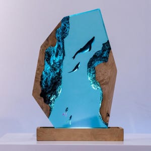 Könnte beinhalten: Dekorative Kunstharzskulptur mit einer Unterwasserszene mit Walen und Tauchern. Das Kunstwerk ist in einer transparenten, geometrischen Form mit Holzseiten und einem Holzsockel eingeschlossen. Die Wale und Taucher sind schwarz, und das Wasser ist hellblau.