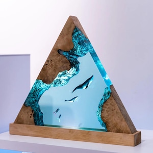 Könnte beinhalten: Eine dreieckige Holzskulptur mit einer Meereslandschaft. Die Skulptur zeigt einen hellblauen Harz-Ozean mit Walen und Tauchern. Der Holzrahmen hat eine natürliche Holzmaserung. Der Sockel ist ein rechteckiger Holzblock.