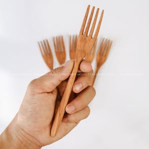 May include: A set of five light brown wooden forks with four tines each, featuring a smooth, natural grain texture. The forks are arranged in a fan-like pattern, with one fork held in the foreground and four others positioned behind it.