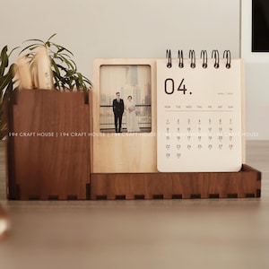Op de afbeelding: Houten bureau-organizer met een kalender voor april en een fotolijst. De organizer is gemaakt van donker hout en heeft een pennenhouder. De fotolijst toont een trouwfoto. De tekst "194 CRAFT HOUSE" is zichtbaar.