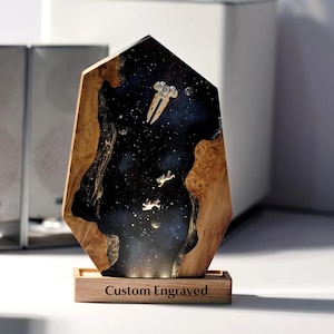 May include: A custom engraved wooden art piece featuring a space scene with a spaceship and astronauts. The artwork is set within a dark, starry background, framed by natural wood. The base of the piece is engraved with the words "Custom Engraved".