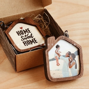 以下が含まれることがあります： 家の形をした木製のキーホルダーが2つ。1つのキーホルダーには「Home Sweet Home」という文字と小さな赤いハートが刻まれています。もう1つのキーホルダーには、ビーチで3人家族の写真が飾られています。