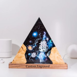 May include: A triangular resin art piece featuring a space scene with astronauts on a moon. The artwork is set in a wooden base with the text "Custom Engraved". The background is a dark blue with stars and galaxies.