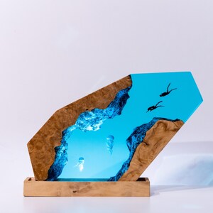 Könnte beinhalten: Dekorative Holz-Kunstwerk mit einer blauen Harz-Ozeanszene mit Quallen und Tauchern. Das Kunstwerk hat einen Holzsockel und eine einzigartige, abgewinkelte Form. Das Holz hat eine natürliche, braune Maserung.