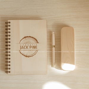 Pode incluir: Um caderno de madeira com encadernação em espiral e um logotipo gravado a laser que diz "CROSSFIT JACK PINE". O caderno está ao lado de uma caneta de madeira e um porta-lápis de madeira.