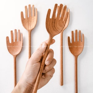 May include: Set of four wooden hand-shaped serving spoons. The spoons are made of natural wood and have a hand-shaped design with five prongs. The spoons are arranged in a row on a white background.