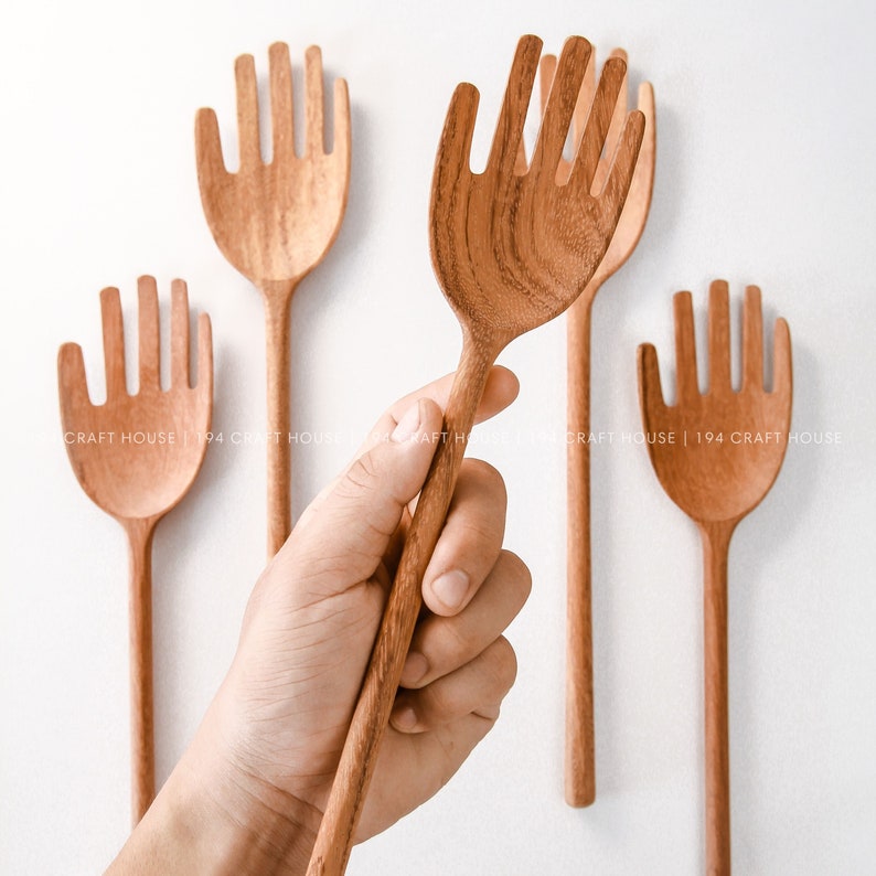 May include: Set of four wooden hand-shaped serving spoons. The spoons are made of natural wood and have a hand-shaped design with five prongs. The spoons are arranged in a row on a white background.
