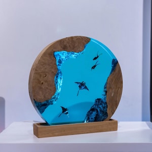 Könnte beinhalten: Ein rundes Holzkunstwerk mit einem türkisfarbenen Harzmittelteil, das Mantarochen-Silhouetten zeigt. Der Holzrahmen hat eine natürliche Maserung, und das Harz leuchtet blau. Es steht auf einem rechteckigen Holzsockel.