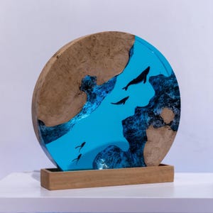 Könnte beinhalten: Runde Holzskulptur mit einem blauen Harzfluss-Design. Die Skulptur hat einen Holzsockel und einen kreisförmigen Holzrahmen mit einem blauen Harzmittelteil. Schwarze Wal-Silhouetten schwimmen im blauen Harzfluss.