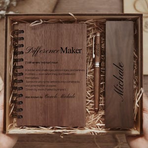 May include: A wooden gift set featuring a notebook, pen, and bookmark, all with a dark wood grain finish. The notebook reads "Difference Maker" with a definition. The pen and bookmark are engraved with the name "Michale". The set is presented in a wooden box.