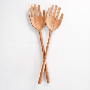 May include: A pair of wooden salad servers shaped like hands. The servers are crossed over each other and are made of light brown wood.