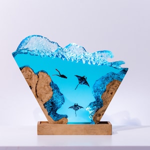 Könnte beinhalten: Dekorative Harzskulptur, die eine Unterwasserszene mit Mantarochen und einem Taucher darstellt. Die Skulptur zeigt einen blauen Ozean, Holzelemente und eine wellenförmige Oberseite. Der Sockel hat eine hellbraune rechteckige Form.