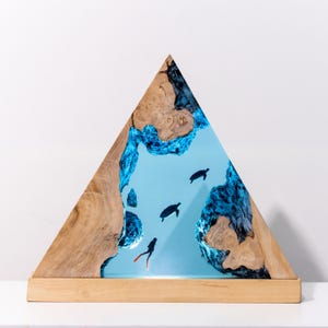 Könnte beinhalten: Eine dreieckige Holzskulptur mit einem hellblauen Harzzentrum, das eine Unterwasserszene darstellt. Die Szene zeigt Meeresschildkröten und einen Taucher. Der Holzrahmen hat eine natürliche Holzmaserung, und die Basis besteht aus hellem Holz.
