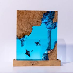 Könnte beinhalten: Holzkunstwerk mit einer blauen Harzszene, die zwei Mantarochen und eine klassische Säule darstellt. Das Kunstwerk ist von einem hellbraunen Holzrahmen umgeben und steht auf einem Holzsockel.