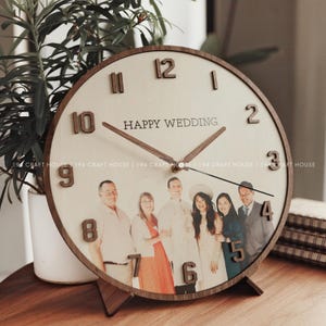 Op de afbeelding: Houten klok met een lichtgekleurde wijzerplaat, een familiefoto en de tekst "HAPPY WEDDING". De klok heeft bruine houten cijfers en wijzers. De klok staat op een houten standaard. Diameter ongeveer 25 cm.
