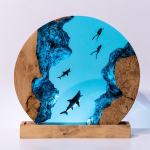 Op de afbeelding: Ronde houten sculptuur met een blauw hars middenstuk dat een onderwater scène uitbeeldt. De scène bevat haaien en duikers. Het houten frame heeft een natuurlijke houtnerf en is gemonteerd op een houten voet.