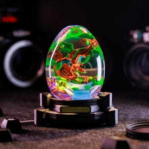Może przedstawiać: Przezroczysta rzeźba z żywicy w kształcie jaja z miniaturową sceną fantasy. Wewnątrz czerwony smok spoczywa w pobliżu wodospadu i drzew. Rzeźba stoi na czarnej, geometrycznej podstawie.