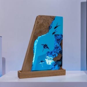 Könnte beinhalten: Holzskulptur mit blauem Harzdesign, das Mantarochen darstellt. Das Kunstwerk hat einen natürlichen Holzrahmen und -sockel. Das blaue Harz erzeugt eine Unterwasserszene, mit schwarzen Mantarochen-Silhouetten.