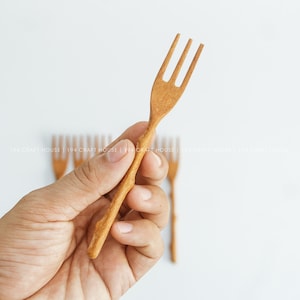 Könnte beinhalten: Eine hellbraune Holzgabel mit einem dreizinkigen Kopf und einem Griff in Form eines natürlichen Zweigs. Die Gabel weist eine strukturierte, organische Oberfläche auf. Weitere identische Holzgabeln sind im unscharfen Hintergrund zu sehen.