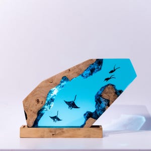 Könnte beinhalten: Ein dekoratives Holzkunstwerk mit einer lebendigen blauen Flusslandschaft aus Harz mit Mantarochen-Silhouetten. Der Holzrahmen hat eine natürliche Holzmaserung. Das Kunstwerk steht auf einem rechteckigen Holzsockel.