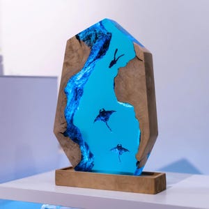 Könnte beinhalten: Dekorative Holzskulptur mit einer blauen Flusslandschaft aus Kunstharz, die Mantarochen und einen Taucher zeigt. Die Skulptur steht auf einem Holzsockel und hat eine einzigartige, geometrische Form. Das Holz hat eine natürliche, hellbraune Farbe.