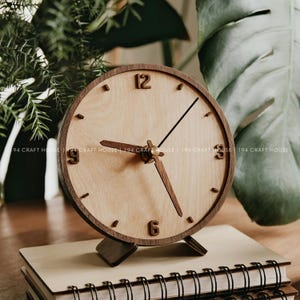 Op de afbeelding: Een houten bureauklok met een licht houten wijzerplaat en donkerbruine rand en wijzers. De klok heeft een standaard en staat op een notitieboek. De cijfers 12, 3, 6 en 9 zijn zichtbaar. De klok heeft een diameter van ongeveer 15 cm.
