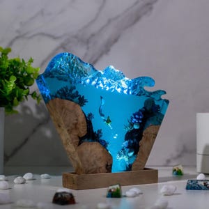 Könnte beinhalten: Dekorative Harzlampe mit einer Unterwasserszene mit Taucher, Korallen und Blasen. Die Lampe hat einen Holzsockel und eine blaue, wellenförmige Oberseite, die ein sanftes Leuchten aussendet. Ein einzigartiger Einrichtungsgegenstand.