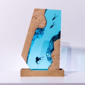 Könnte beinhalten: Dekorative Holzskulptur mit einer hellblauen Flusslandschaft aus Kunstharz, die Taucher und einen Oktopus zeigt. Das Holz hat eine natürliche Maserung und ist hellbraun. Die Skulptur ist auf einem Holzsockel montiert.
