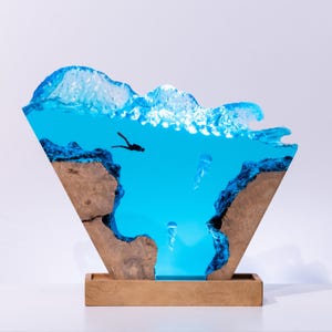 Könnte beinhalten: Dekorative Harzskulptur, die eine Unterwasserszene darstellt. Das Kunstwerk zeigt einen Taucher, Quallen und eine Welle. Die Skulptur ist in einem Holzrahmen und Sockel gesetzt, mit einem leuchtend blauen Ozean und detaillierten Felsformationen.