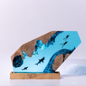 Könnte beinhalten: Dekorative Holzskulptur mit einer blauen Harz-Ozeanszene mit Haien und Tauchern. Die Skulptur hat einen Holzsockel und eine einzigartige Form. Das blaue Harz simuliert Wasser, in dem Haie und Taucher schwimmen.