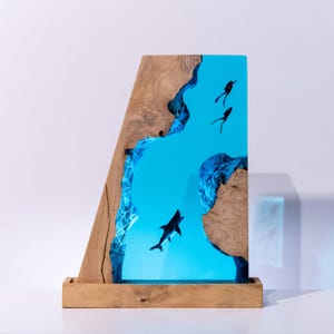 Könnte beinhalten: Holzskulptur mit einer Szene unter Wasser, mit einem Hai und Tauchern in einem blauen Harzfluss. Der Holzrahmen hat eine natürliche Maserung. Der Harzfluss wird mit blauem Licht beleuchtet, was einen Unterwasser-Effekt erzeugt.