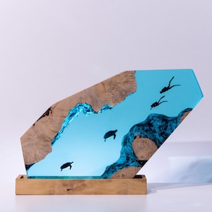 Könnte beinhalten: Dekorative Holzskulptur mit einer blauen Harz-Ozeanszene mit Schildkröten und Tauchern. Die Skulptur hat einen Holzsockel und ein einzigartiges, abgewinkeltes Design. Das Holz hat eine natürliche, hellbraune Farbe und das Harz ist ein leuchtendes Blau.