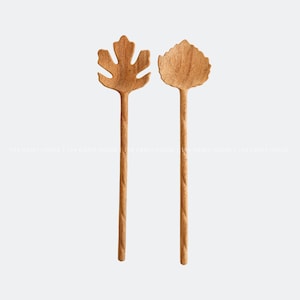 Può includere: Due cucchiai da portata in legno con teste a forma di foglia. I cucchiai sono realizzati in legno naturale e hanno un aspetto rustico e artigianale.