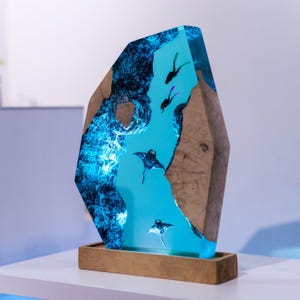 Könnte beinhalten: Dekorative Resin-Skulptur mit blauem Innenraum, die Mantarochen und Taucher darstellt. Die Skulptur ist in einem Holzrahmen eingefasst und steht auf einem Holzsockel. Der blaue Innenraum hat einen strukturierten, leuchtenden Effekt.