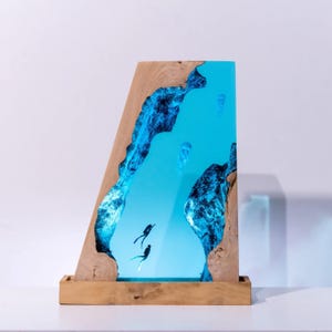 Könnte beinhalten: Ein Holz- und Kunstharz-Kunstwerk mit einer Unterwasserszene mit Tauchern und Quallen. Das Kunstwerk ist in einem Holzrahmen und Sockel eingefasst. Das Kunstharz ist leuchtend blau, mit Tauchern und Quallen, die darin schweben.