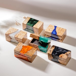 May include: A collection of handcrafted wooden ring boxes with colorful epoxy resin accents. The boxes feature a natural wood grain and are filled with green, blue, orange, white, and black resin. One box is open, revealing a ring.