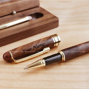 Può includere: Una penna in legno con dettagli dorati e una cartuccia d'inchiostro nera. La penna è incisa con il testo "Your Name".