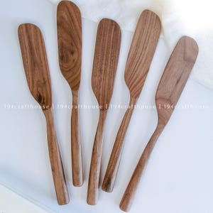May include: Five wooden butter spreaders with a natural brown grain pattern and smooth, rounded edges. Each spreader features a long, slender handle and a wide, flat blade, arranged in a fan-like formation on a plain white surface.
