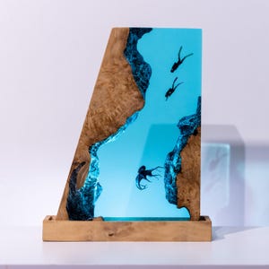 Könnte beinhalten: Dekorative Harzskulptur mit einer Unterwasserszene mit einem Oktopus und zwei Tauchern. Die Skulptur ist von Naturholz mit hellbraunem Finish umrahmt und steht auf einem Holzsockel. Die Meereslandschaft wird blau beleuchtet.