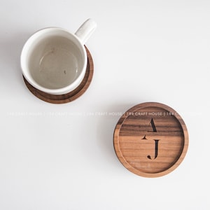 May include: Two round wooden coasters with a dark brown grain. One coaster supports a white ceramic mug, while the other features the engraved black letters AJ in the center. The text 194 CRAFT HOUSE is repeated across the image.