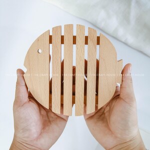 Könnte beinhalten: Ein Holz-Untersetzer in Fischform mit einem Lamellendesign. Der Untersetzer ist hellbraun und wird von zwei Händen gehalten.