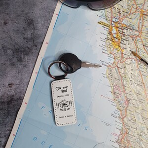 Peut inclure: Un porte-clés en cuir blanc avec des coutures noires et le texte "On the Road" en noir. Le porte-clés présente également une petite illustration noir et blanc d'un camping-car devant des montagnes. Le texte "Depuis 2020" et "Lucas & Senoit" sont également imprimés sur le porte-clés.